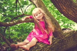 Playful girl in a tree, unafraid of mistakes or criticism, with green leaves and sunshine behind her